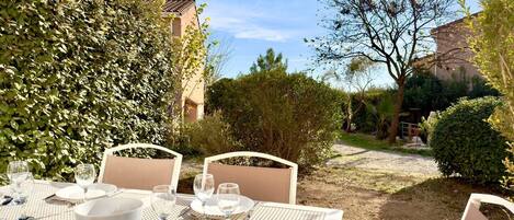An outdoor dining area with white tables and chairs, surrounded by greenery and a gravel path.