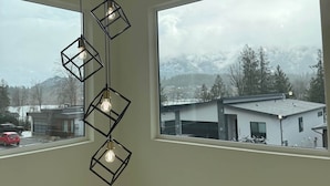 A modern interior with a geometric pendant light fixture and large windows offering a view of a snowy landscape and buildings. - Lakeside Living – New 4 Bedroom Retreat (Lake Cowichan)