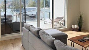 A spacious living room with a grey sectional sofa, a wooden coffee table, and a view of a deck and mountains through large sliding glass doors. - Lakeside Living – New 4 Bedroom Retreat (Lake Cowichan)