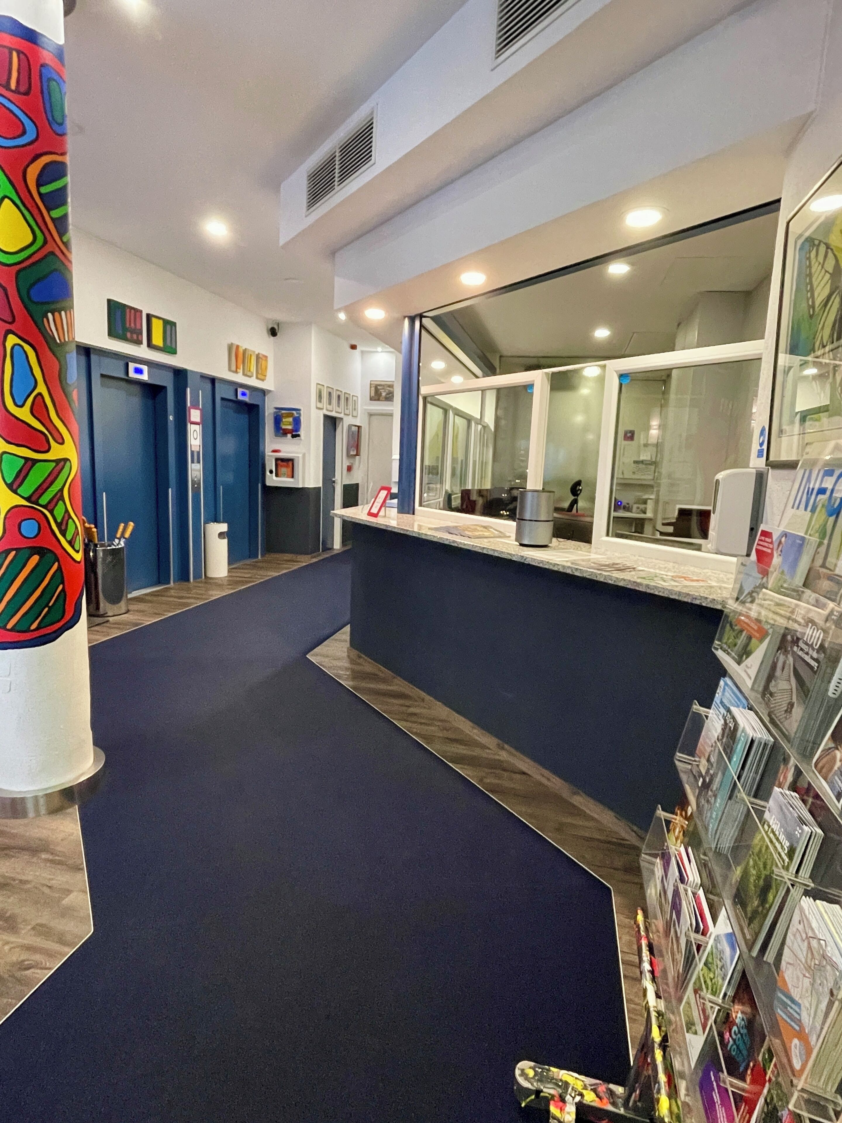 Un vestíbulo de hotel con una recepción, un pilar colorido y un expositor de revistas.
