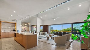 A modern kitchen with a central island, a dining area with a sofa and chairs, and large windows offering a view of the outdoors.