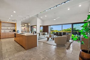 A modern kitchen with a central island, a dining area with a sofa and chairs, and large windows offering a view of the outdoors.