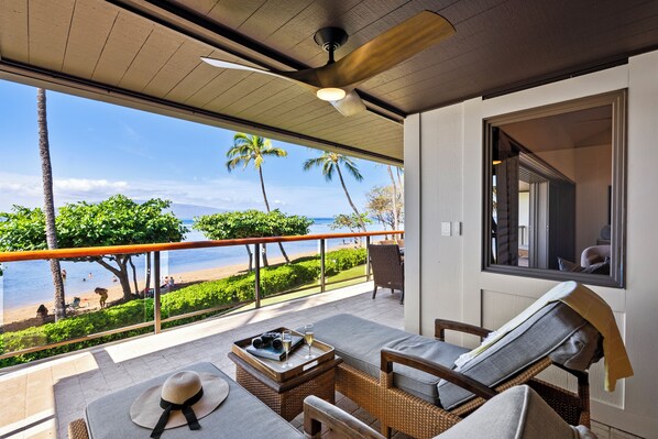 A balcony with a view of the beach, wicker furniture, and a ceiling fan.