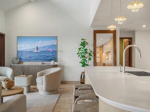 A modern kitchen with a large island, a dining area with chairs, a television displaying a scenic ocean view, and a potted plant.