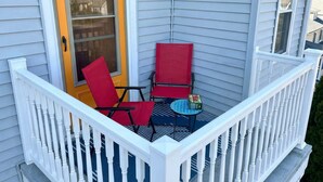 Un balcon d’angle avec une balustrade blanche, deux chaises rouges, une petite table ronde et une porte dotée d’un cadre jaune.