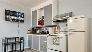 Une cuisine compacte avec un réfrigérateur blanc, une cuisinière et un four à micro-ondes. On y trouve une table à manger avec des chaises, et un téléviseur fixé au mur qui diffuse un service de streaming.