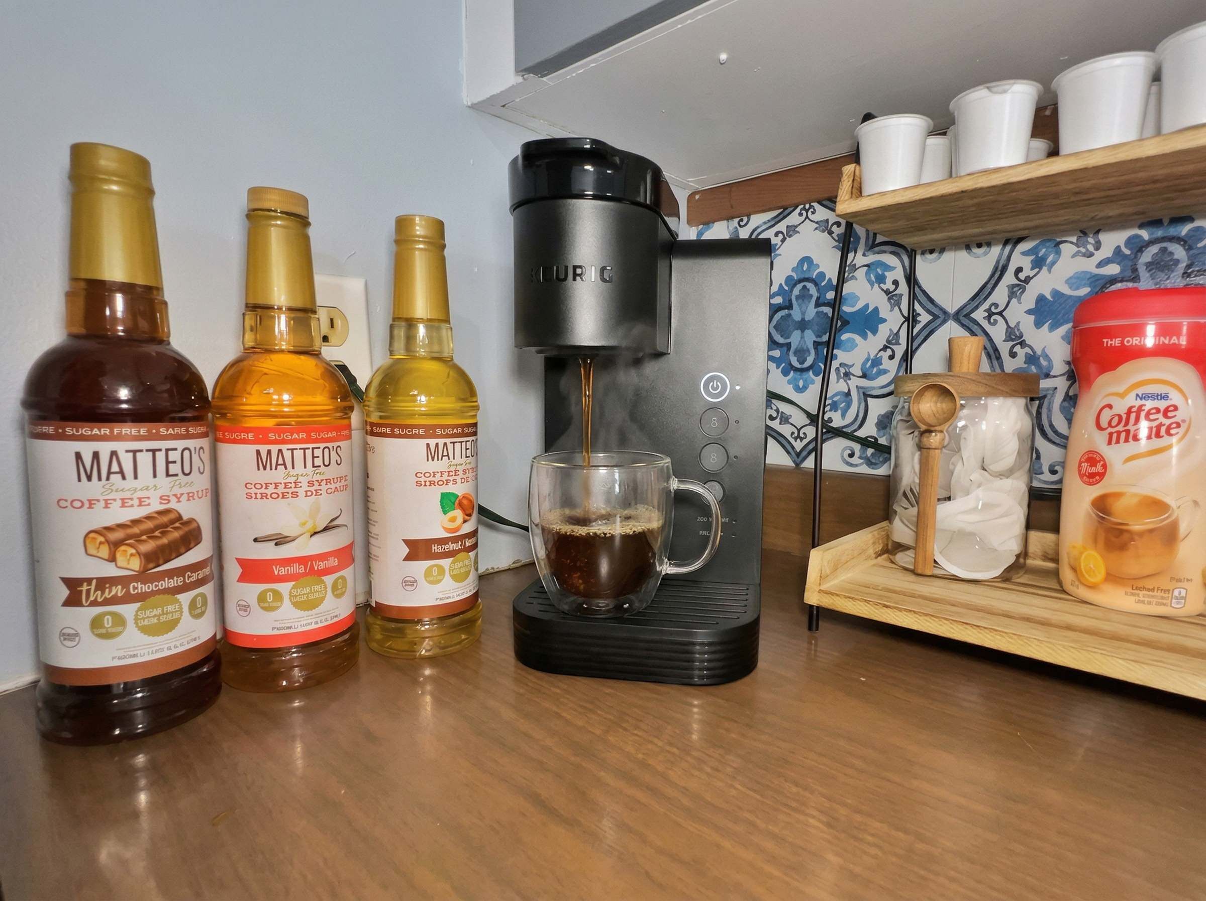 Una encimera de cocina con tres botellas de sirope de café, una cafetera y un cartón de Nestlé Coffee Mute.