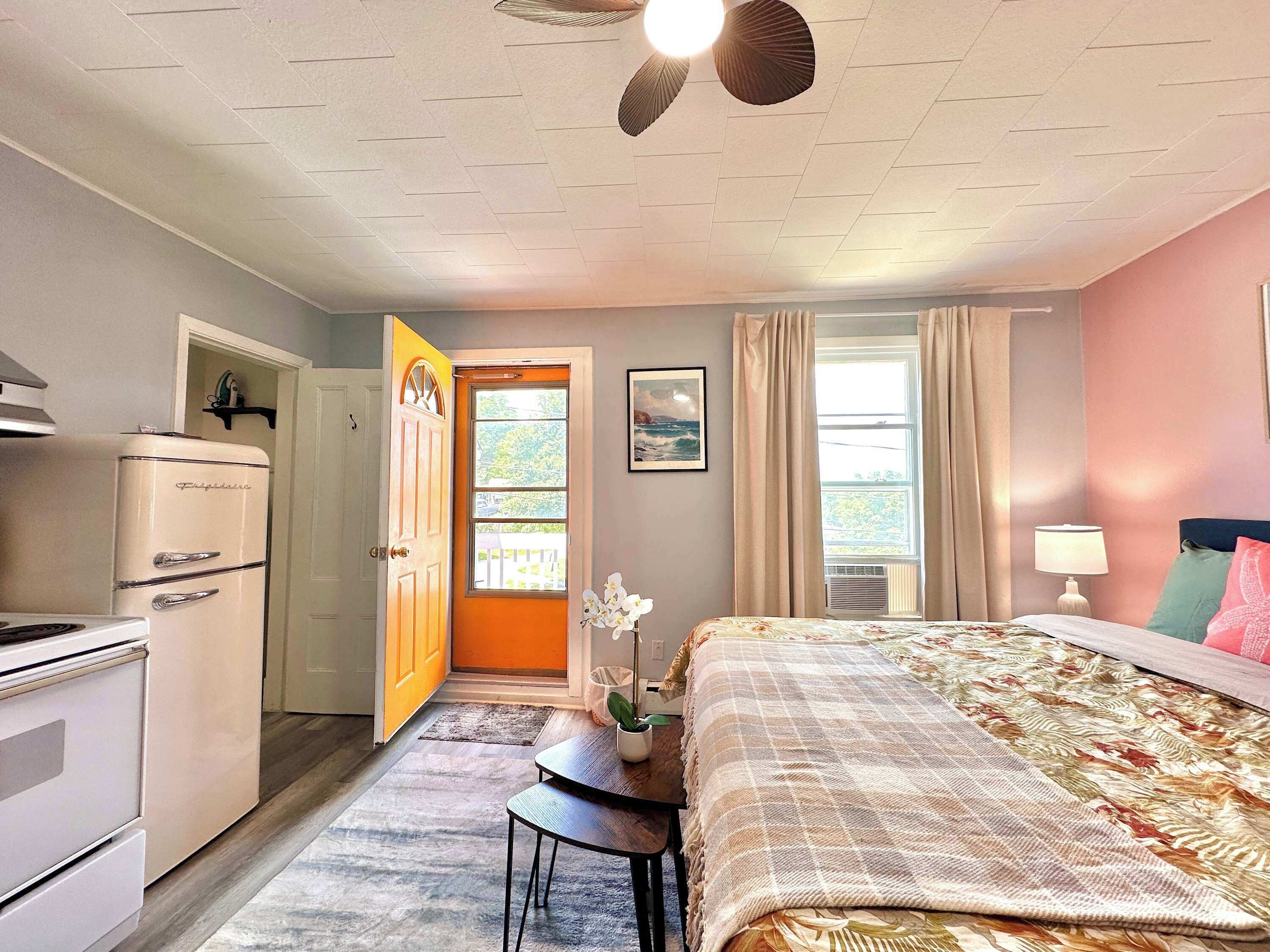 Ein Schlafzimmer mit Bett, Schreibtisch, Kühlschrank, einer Tür zum Balkon und einem mit Vorhängen drapierten Fenster.