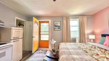 Ein Schlafzimmer mit Bett, Schreibtisch, Kühlschrank, einer Tür zum Balkon und einem mit Vorhängen drapierten Fenster.