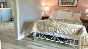 Un dormitorio con una cama, dos lámparas de noche, un cuadro en la pared y una cocina al fondo.