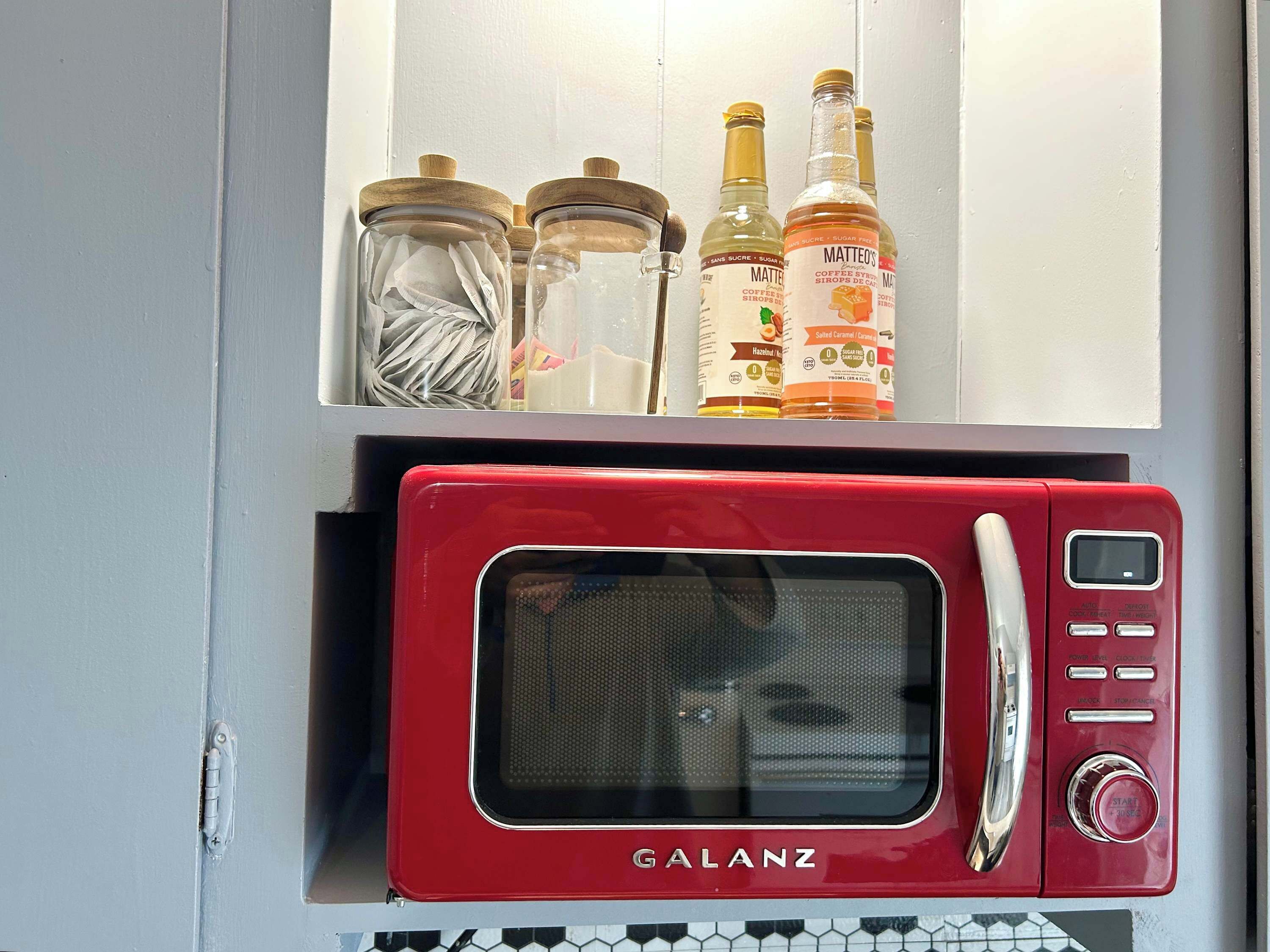 Un four à micro-ondes Galanz rouge, doté d’un écran numérique, d’un panneau de commande et d’un bouton de démarrage. Au-dessus, on trouve trois bocaux en verre avec des couvercles, dont l’un contient un chiffon blanc, et deux bouteilles étiquetées « sirop de café Matteo ».