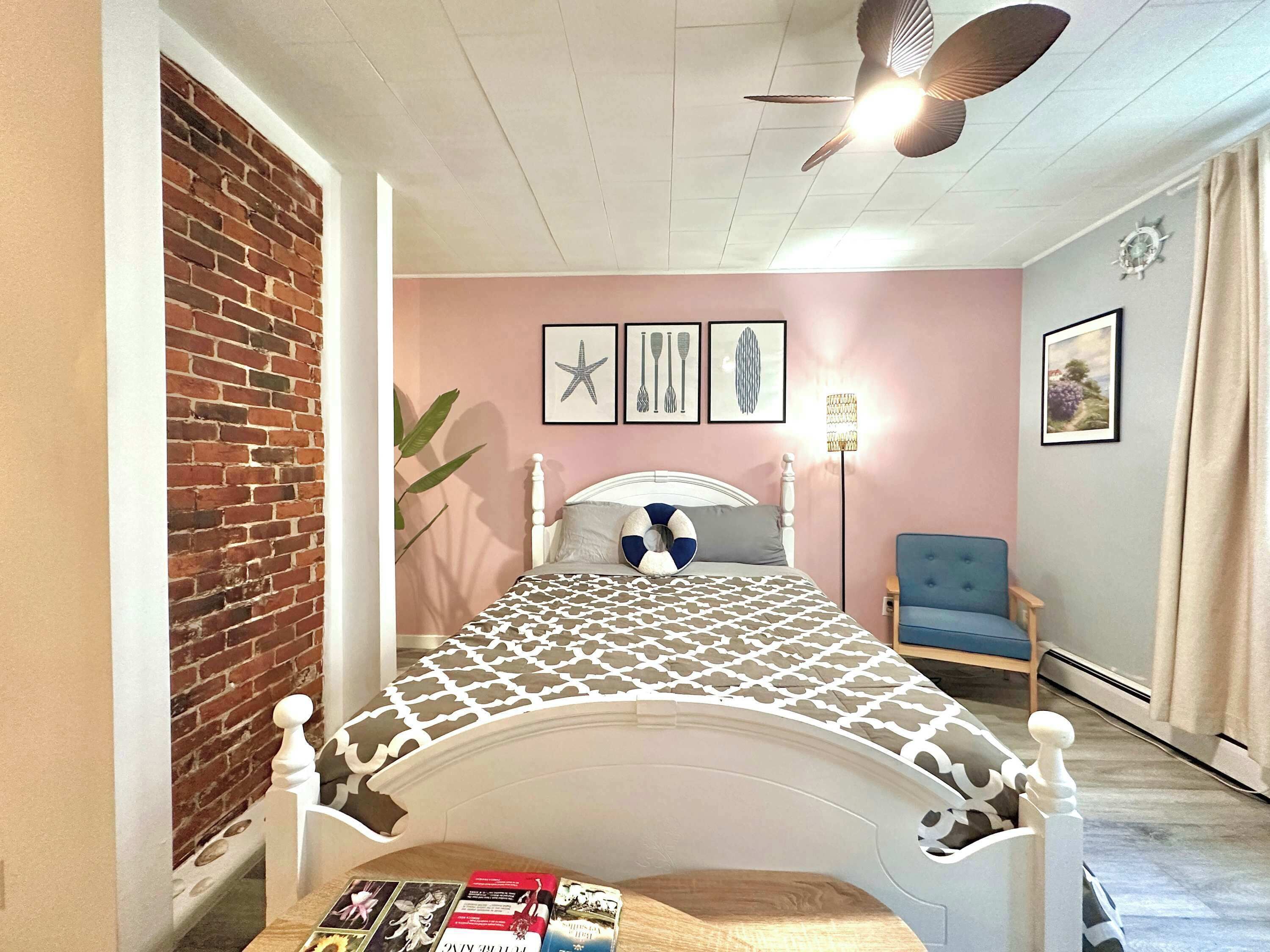 A bedroom with a brick wall, a white bed with a patterned bedspread, a ceiling fan, and a blue chair.