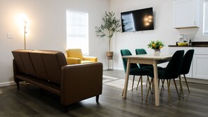 En moderne stue med sofa, spisebord med stole, fjernsyn monteret på væggen og et køkken i baggrunden.