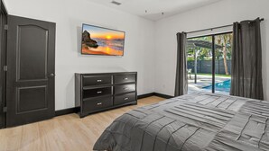 Un dormitorio con cama, cómoda, ventilador de techo, un cuadro y vista a una piscina al aire libre.