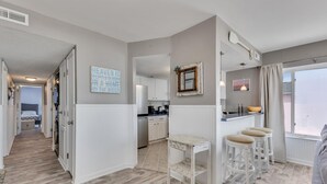 Una cucina moderna con mobili bianchi, un angolo bar con sgabelli, un'ampia finestra vista e un corridoio che conduce a una camera da letto.