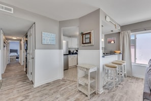 A modern kitchen with white cabinetry, a bar area with stools, a large window with a view, and a hallway leading to a bedroom. - Sunskipper D9 - Oceanfront condo with Private Decks! (Carolina Beach)