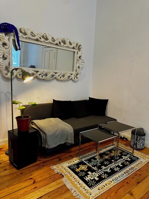 A modern living room with a dark grey sofa, a small black coffee table, a red potted plant, a black floor lamp, and a patterned rug on a wooden floor. - Quiet Altbau 2-Room Apartment with Terrace | Central Berlin near Parks & S-Bahn (Berlin)