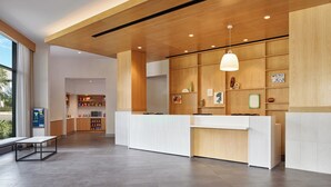 A modern reception area with a wooden reception desk, a hanging pendant light, and a view of the kitchen in the background. - Hyatt House at Ho'opili (Ewa Beach)
