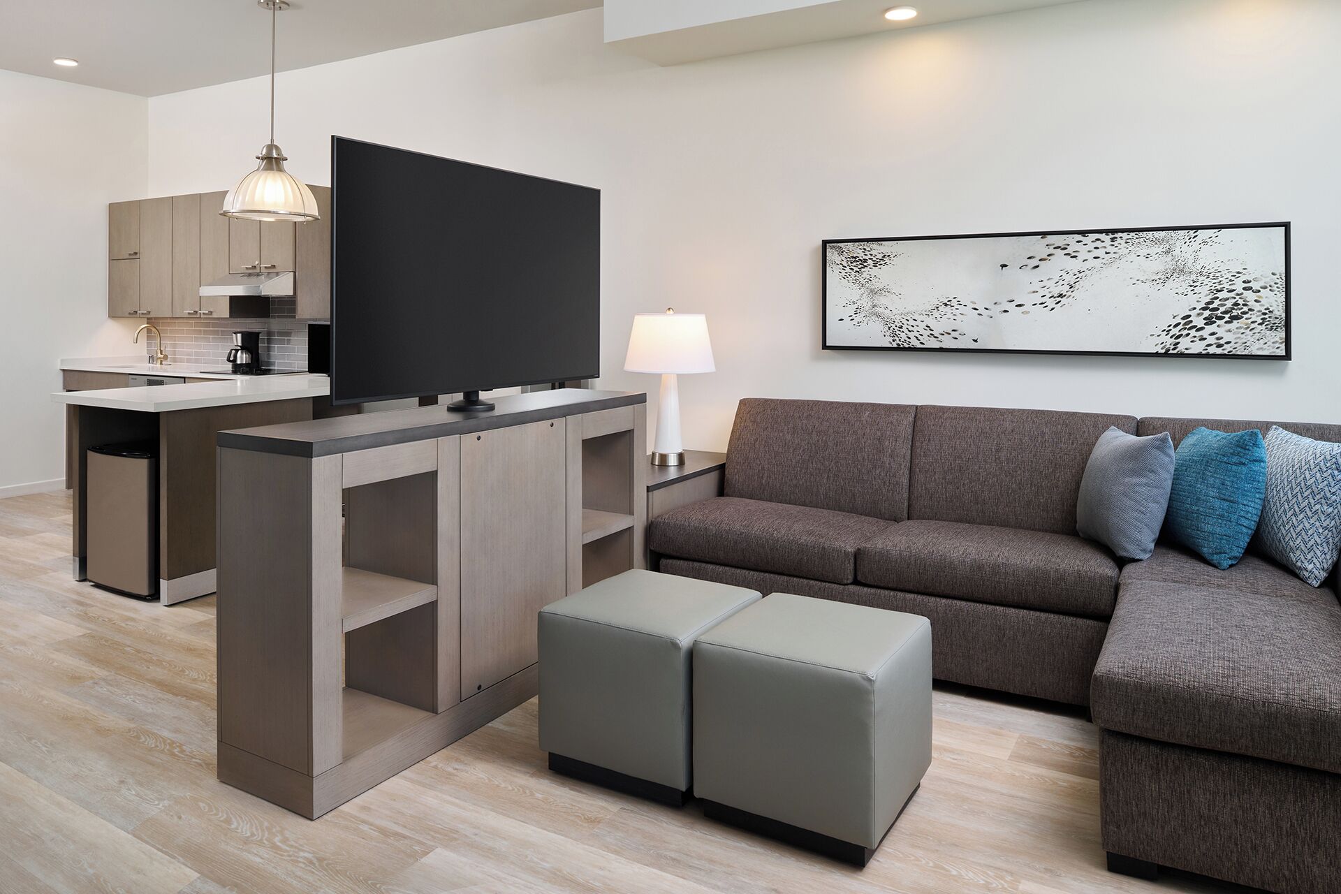 A modern living room with a flat-screen TV, grey sofa, ottomans, a dining area with a kitchen in the background, and a wall-mounted abstract art piece.