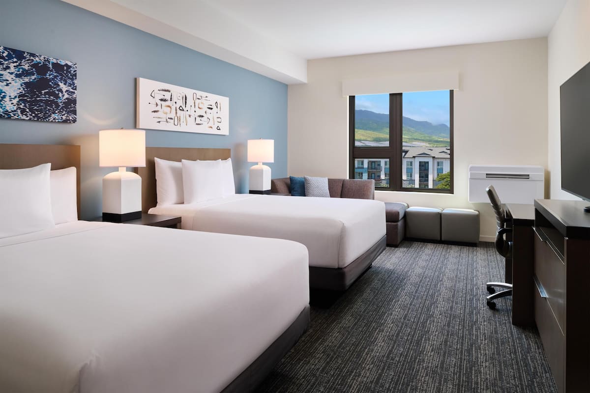 Une chambre d’hôtel avec deux lits, un bureau, une chaise, une tél évision et une fenêtre donnant sur des immeubles et des montagnes.