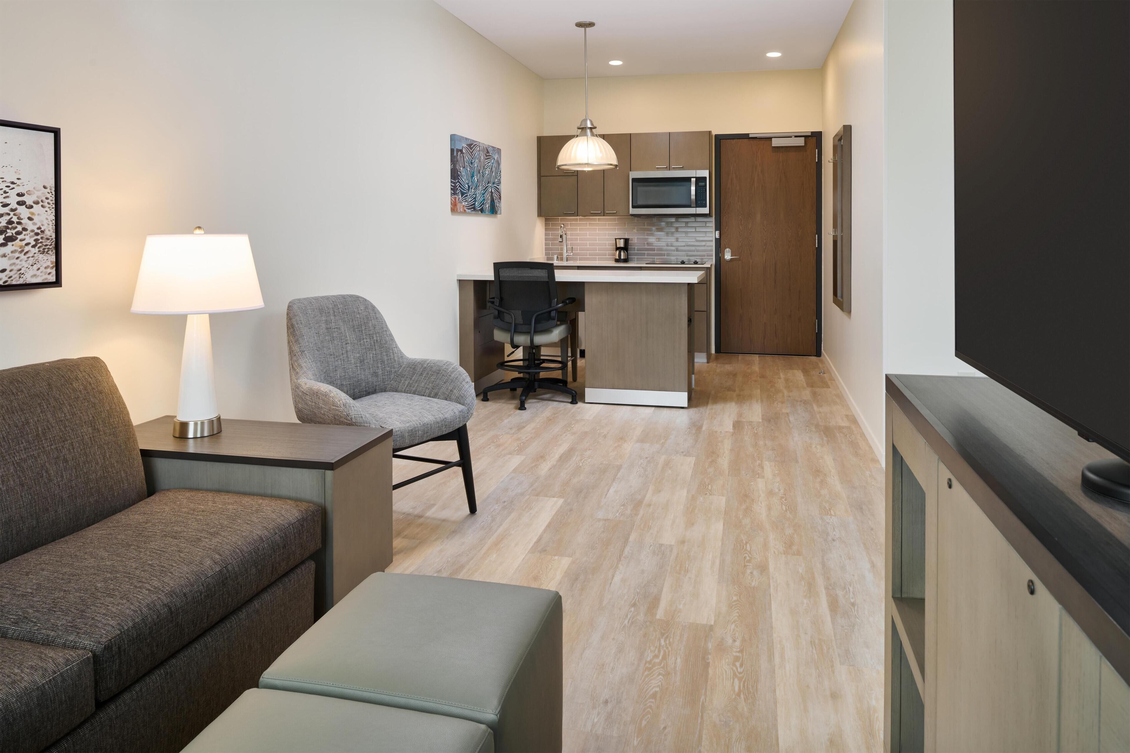 A compact living space with a sofa, a chair, a small table with a lamp, a kitchenette with a microwave, and a TV.