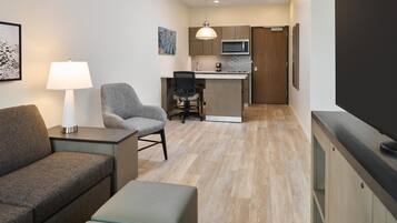 A compact living space with a sofa, a chair, a small table with a lamp, a kitchenette with a microwave, and a TV.