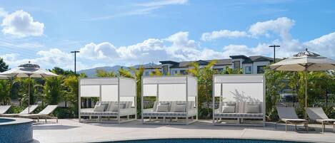 A pool area with lounge chairs and umbrellas, a clear sky, and a residential building in the background.