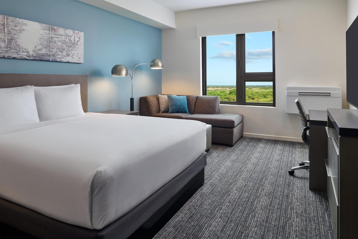 Une chambre d’hôtel avec un grand lit, un bureau, une chaise, un canapé, une fenêtre donnant sur un beau paysage et une œuvre d’art accrochée au mur.