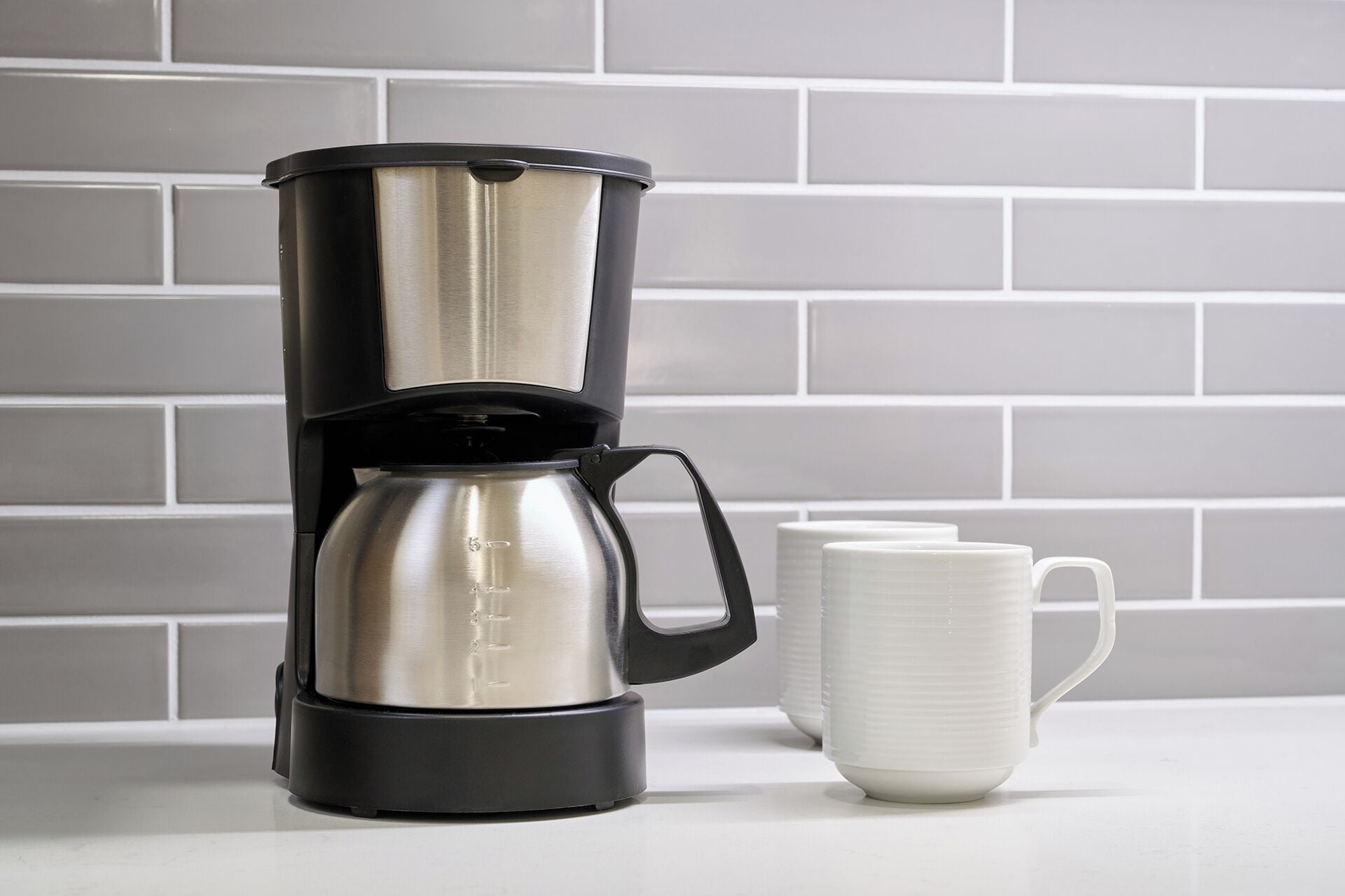 Una cafetera con base negra y jarra de acero inoxidable, una taza blanca y un revoque de azulejos.