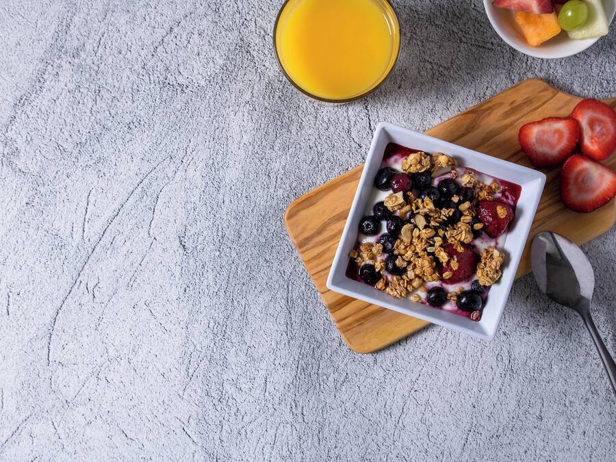 Uma salada de frutas com granola sobre uma tábua de madeira, um copo de suco de laranja e uma tigela com frutas mistas em uma superfície texturizada.