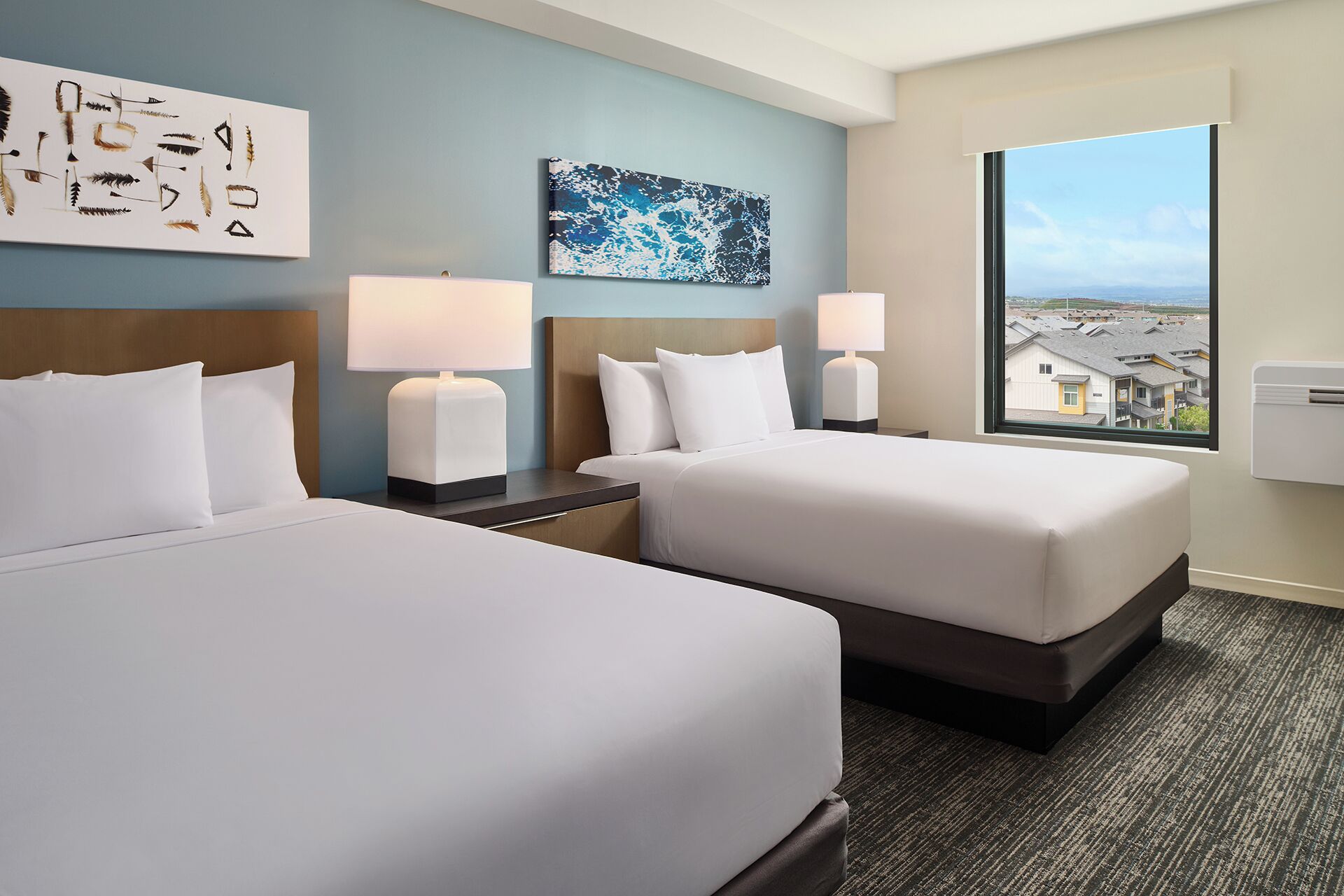 A hotel room with two beds, a large window with a view, a painting on the wall, and a decorative piece above the bed.