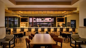 Eine moderne Bar mit einer langen Theke, verschiedenen Sitzgelegenheiten und Blick auf Pool und Palmen.