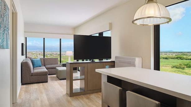 A modern living room with a sofa, TV, and dining area with a view of the countryside. - Hyatt House at Ho'opili (Ewa Beach)