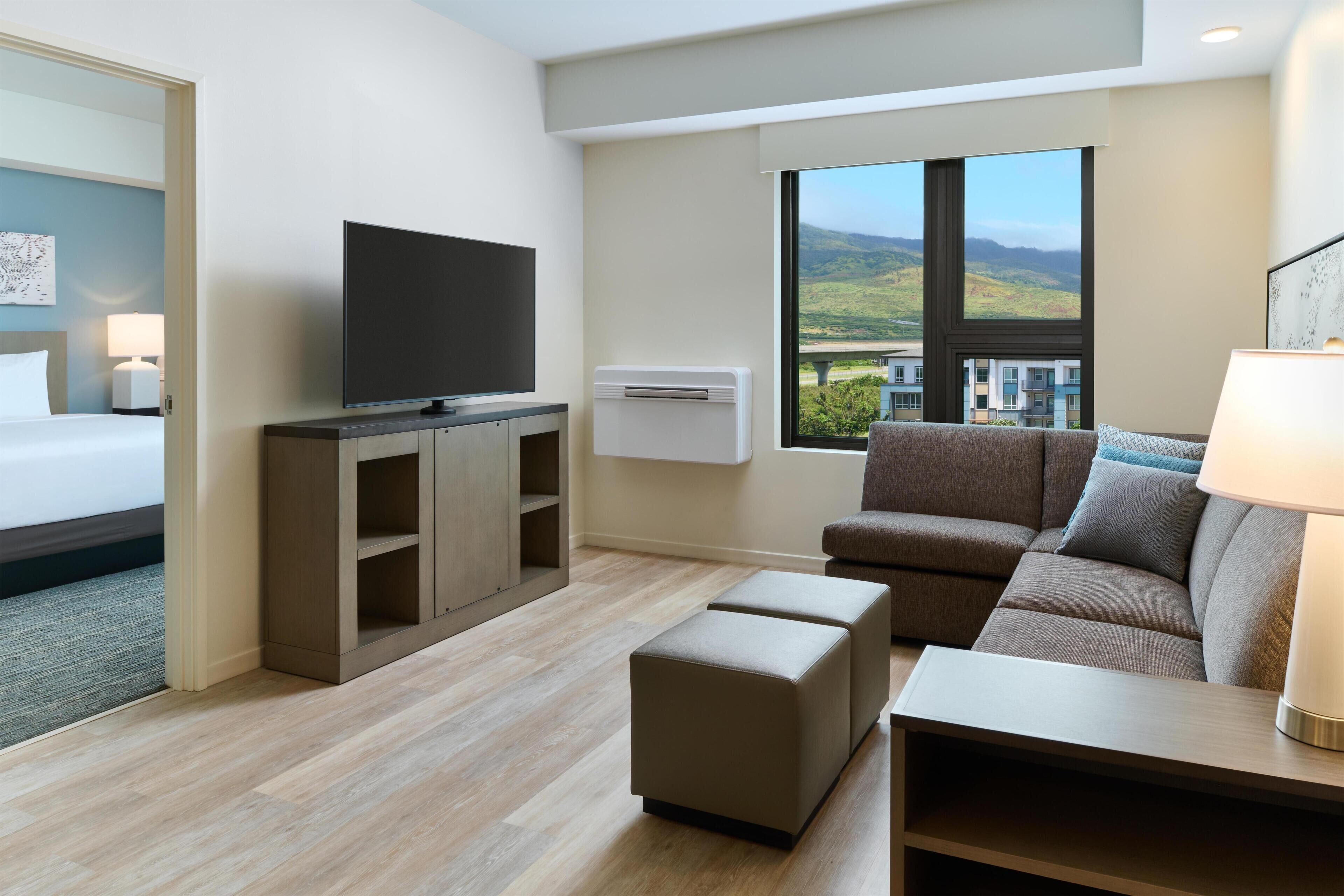Moderni hotellihuone, jossa on taulutelevisio, sohva, sohvapöytä ja suuri ikkuna, josta avautuu näkymä rakennuksiin ja kukkuloille.