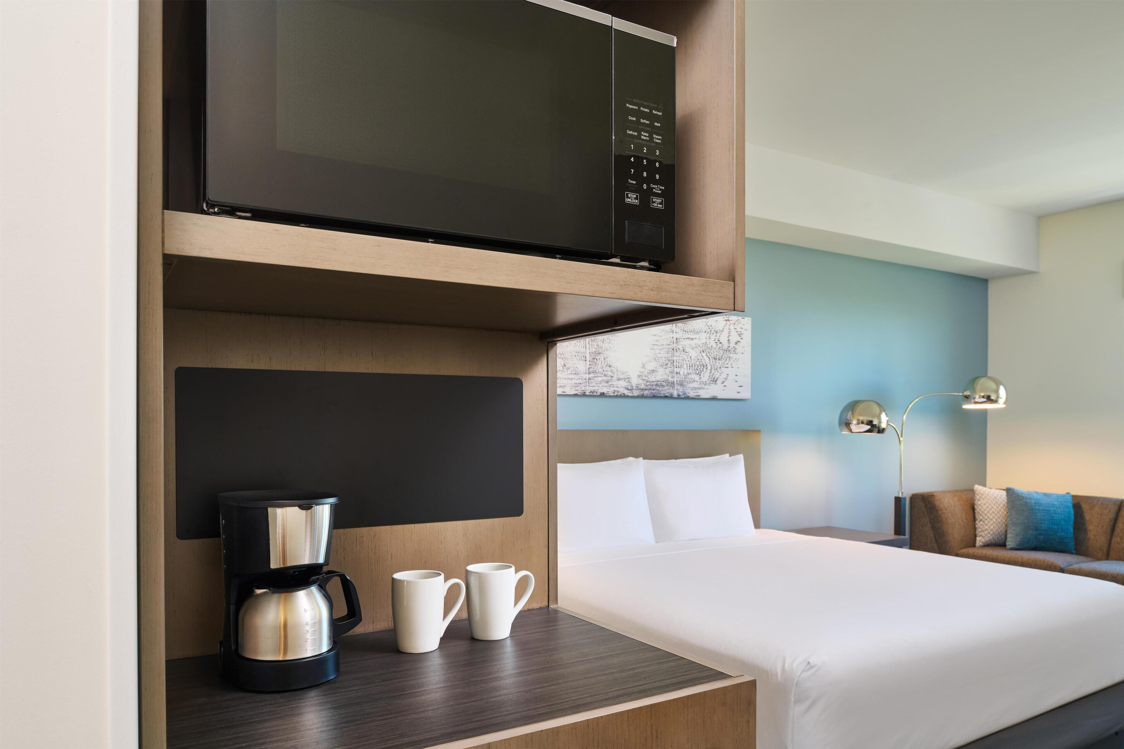 Habitación de hotel con cama, cafetera, dos tazas, microondas y televisor montado en la pared.
