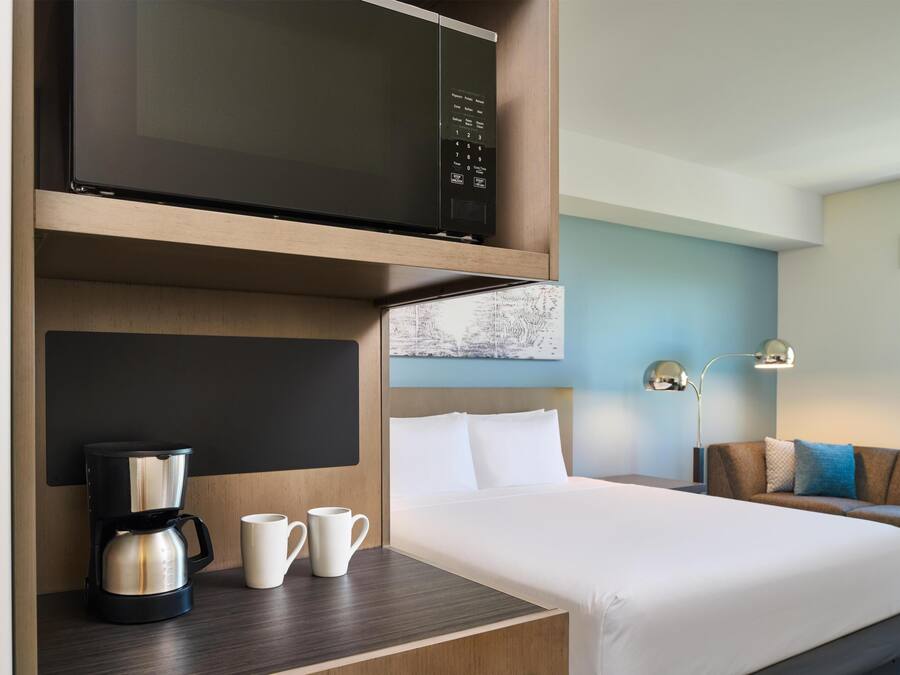 Habitación de hotel con cama, cafetera, dos tazas, microondas y televisor montado en la pared.