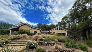 A two-story house with a stone pathway leading to the entrance, surrounded by landscaped gardens and trees. - Oak & Vine: Pool | Hot Tub | Fire Table | BBQ (Glen Ellen)