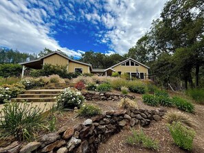 A two-story house with a stone pathway leading to the entrance, surrounded by landscaped gardens and trees. - Oak & Vine: Pool | Hot Tub | Fire Table | BBQ (Glen Ellen)