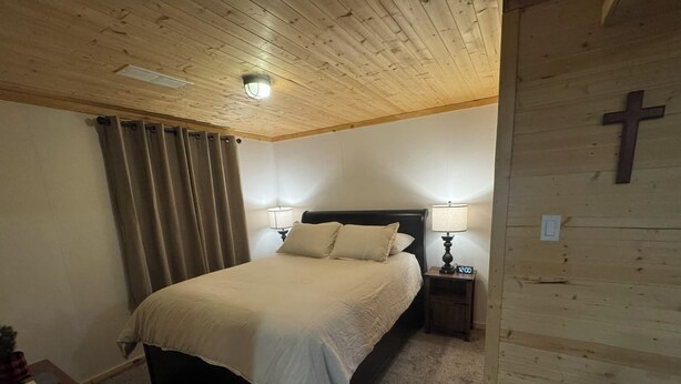 A bedroom with a wooden ceiling, a bed with a beige bedspread, two bedside lamps, and a wooden cross on the wall.