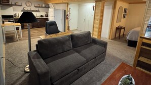 A living room with a grey sofa, a dining table with chairs, a kitchen area, and a wooden ceiling.