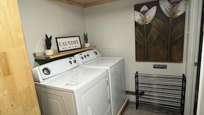 Une buanderie équipée de deux machines à laver et deux sèche-linge blancs, une pancarte indiquant « LAUNDRY WASH, DRY & FOLD » (buanderie, lavage, séchage et pliage), une petite étagère avec des plantes et un tableau représentant des fleurs accroché au mur.