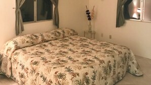 A bedroom with a floral bedspread, two windows with curtains, a small table with a vase, and a wall-mounted light fixture.