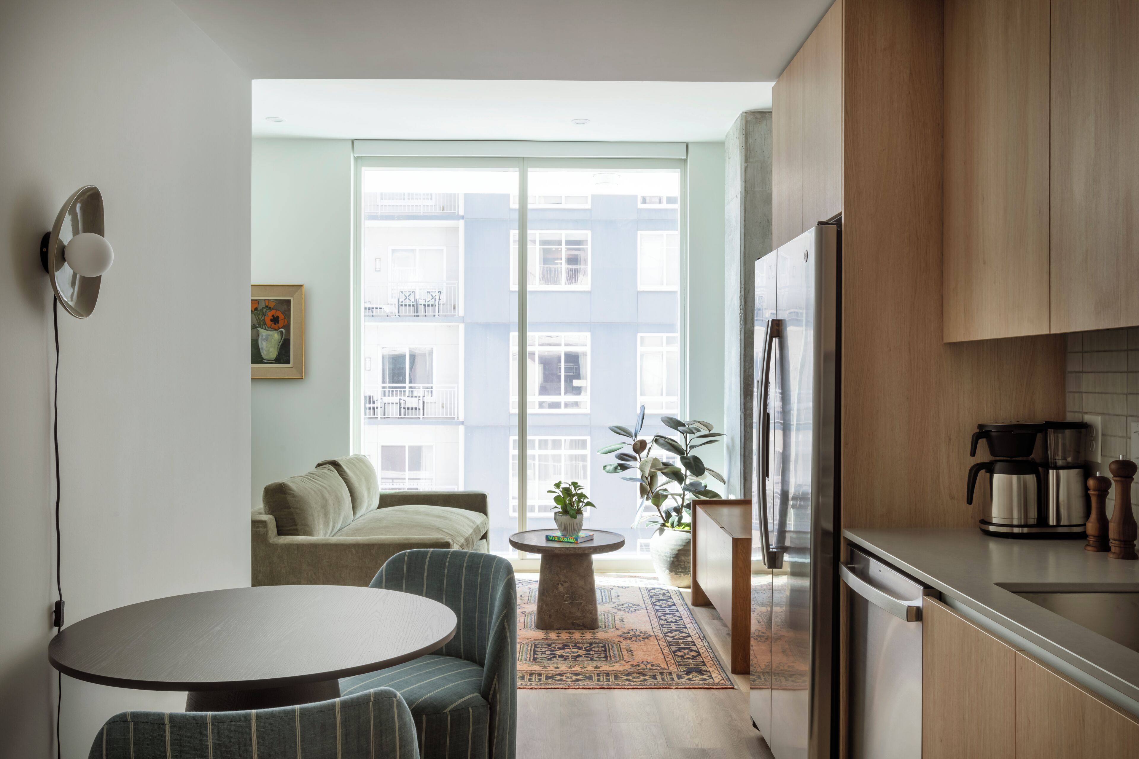 A modern kitchen with a dining area, a refrigerator, a coffee maker, and a view of a building through the window.