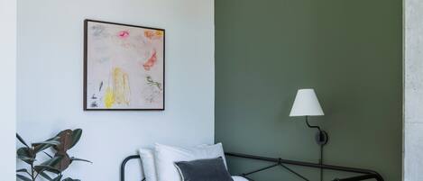 Une chambre à coucher avec un lit, une lampe de chevet, une plante en pot et une œuvre d’art encadrée accrochée au mur.