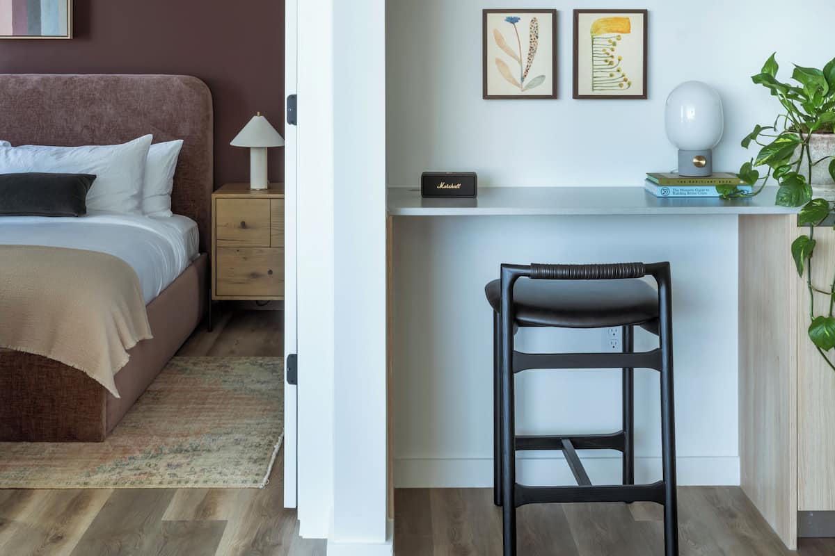 Un dormitorio con cama, mesita de noche, lámpara y un pequeño escritorio con silla y planta.