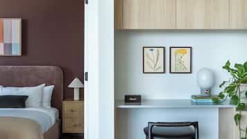 A bedroom with a bed, bedside table, lamp, and a small desk area with a chair and a plant.