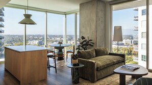 A modern living room with a sofa, a coffee table, a dining area with chairs, and a large window offering a city view. - ROOST Rainey (Austin)