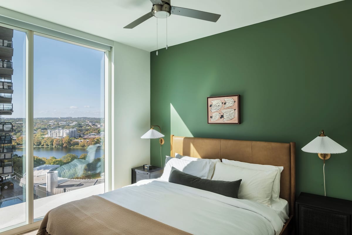 Un dormitorio con un ventanal grande, una cama, un ventilador de techo y vistas a un paisaje urbano.
