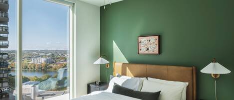 A bedroom with a large window, a bed, a ceiling fan, and a view of a cityscape.