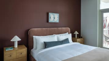 A neatly made bed with white and dark pillows, a brown headboard, and a framed artwork above. There are two bedside tables with lamps and a book on one.